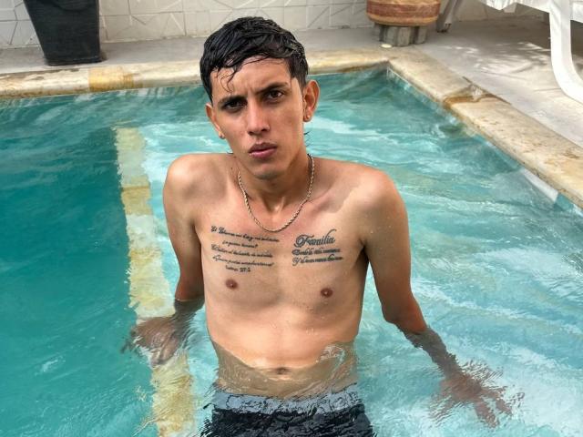 DannaXFabian, shirtless, wet, in a pool, with tattoos on chest, short dark hair, serious expression, small earrings, chain necklace, black swim trunks, standing, water up to waist.
