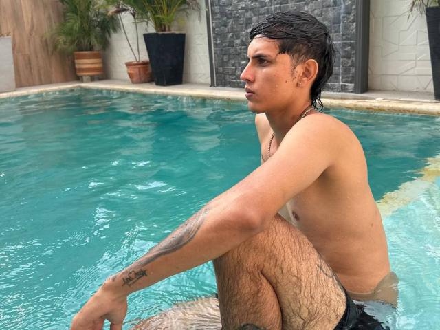 DannaXFabian with wet black hair, shirtless, sitting in a pool, showing a tattoo on his left arm, wearing black swim trunks, looking to the side with a serious expression.
