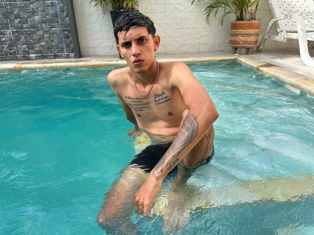 DannaXFabian, shirtless, with tattoos, black wet shorts, necklace, in a pool, leaning forward, looking at camera, water up to thighs, right hand on pool edge.