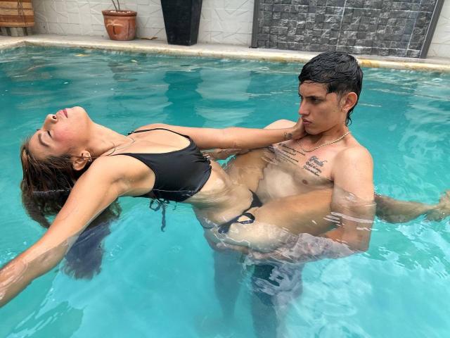 DannaXFabian with tits in a black bikini, arms stretched, head back, being held up in a pool by a shirtless DannaXFabian with a tattoo and a hard-on.