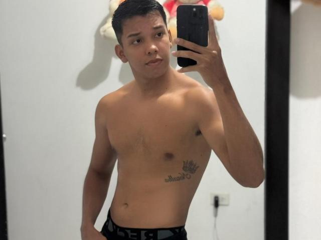 EdwinBlaze, shirtless, holding phone, mirror selfie, black pants, crown tattoo on right side, short dark hair, light skin, slight smile, standing, room with white wall.