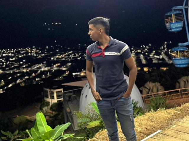 EdwinBlaze stands with hands in jeans pockets, wearing a dark blue polo with red stripe and white logo, looking to the side, in a night-time city view.