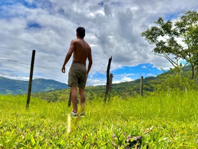 EdwinBlaze stands shirtless in green grass, facing away, wearing khaki shorts and white sneakers, with a fence and cloudy sky behind him, on a hill with trees.