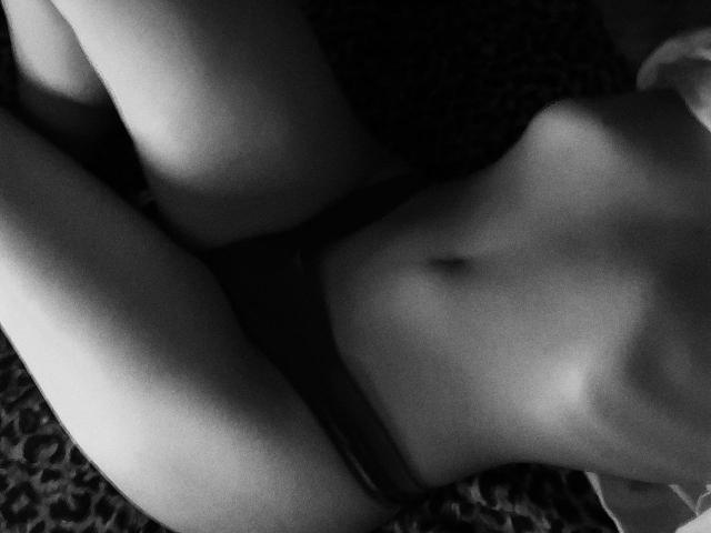 Black and white photo of AngelsBoobsX lying down, showing her flat stomach, small nipples, and dark panties, with her legs slightly apart. Her pussy is mostly covered.