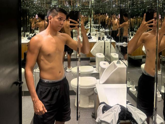 EdwinBlaze, shirtless, with a toned, muscular, tan body, black underwear, and short black hair, taking a mirror selfie in a bathroom with multiple toilets and mirrors.