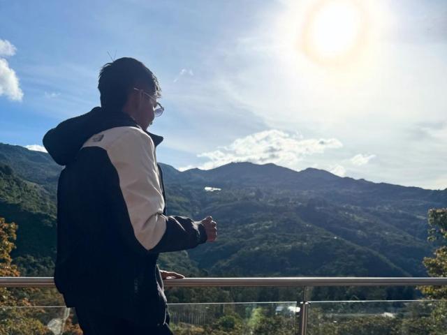 EdwinBlaze with glasses, black and white jacket, standing on metal railing, looking at sunlit mountains, holding camera, blue sky, sun flare, green hills in background, day.