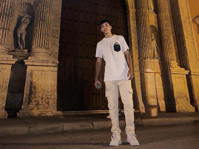 EdwinBlaze stands in front of a dark wooden door, wearing a white tee, beige pants, and white sneakers, holding a can in his right hand, looking up.