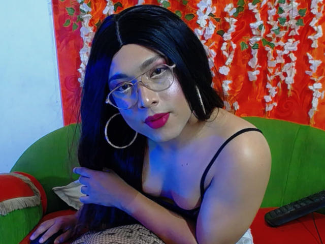MelisaColins with long black hair, glasses, hoop earrings, red lipstick, black bra, blue lighting, sitting on green couch, hands on lap, remote control on right. Boobs visible.
