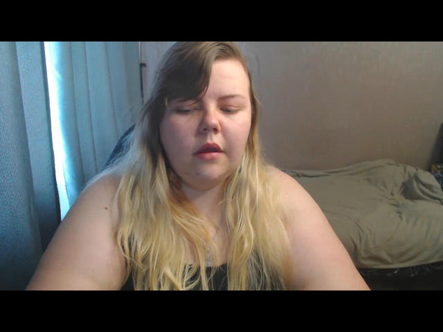 AsmiTKeyn has a chubby face with blonde, slightly messy hair, pale skin, and red lips, sitting with arms forward, wearing a grey top, looking down, in a bedroom.