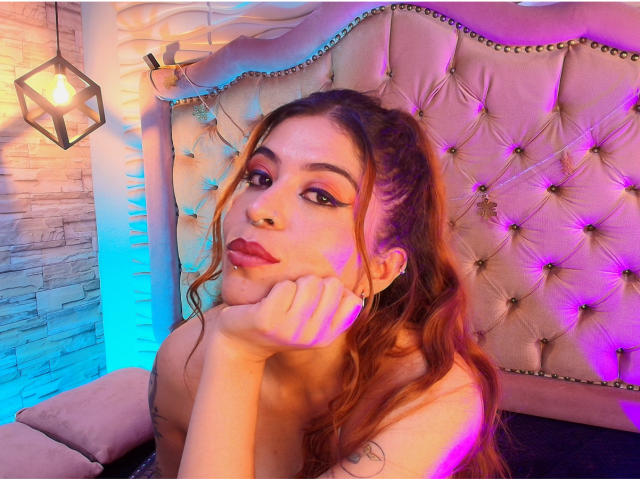 AlaiaLove, com cabelos ruivos ondulados, sombra rosa e batom rosa, apoiando o queixo na mão, exibindo seus seios e um piercing nasal fofo, em um quarto iluminado em tons de roxo..