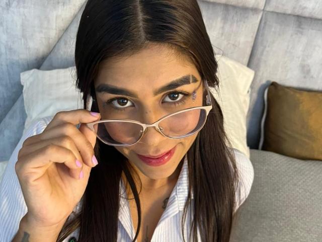 VioletaGates with long brown hair, wearing glasses, smiling, holding them with her hand, pink nails, red lipstick, tattoo above right eyebrow, white shirt, grey headboard.