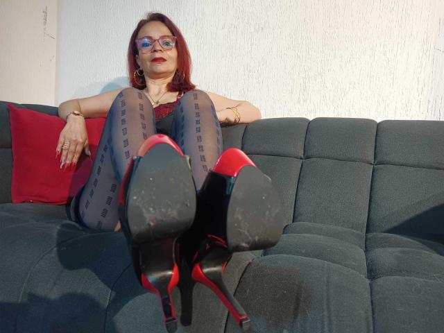 MissEllien with red hair, glasses, and red lipstick, wearing black sheer tights and red high heels with black soles, sitting on a green couch, legs up, showing heels.