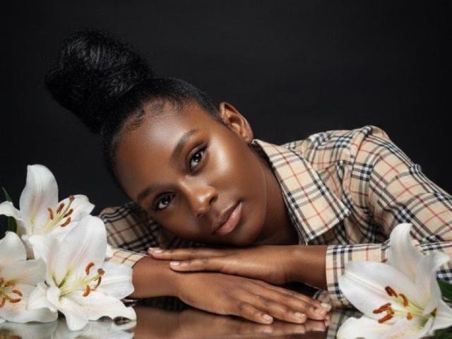 SelinaTae, à la peau foncée, avec des cheveux noirs attachés en chignon, portant une chemise à carreaux, reposant la tête et les mains sur des lys blancs aux étamines brunes. Ses yeux sont doux et sérieux..