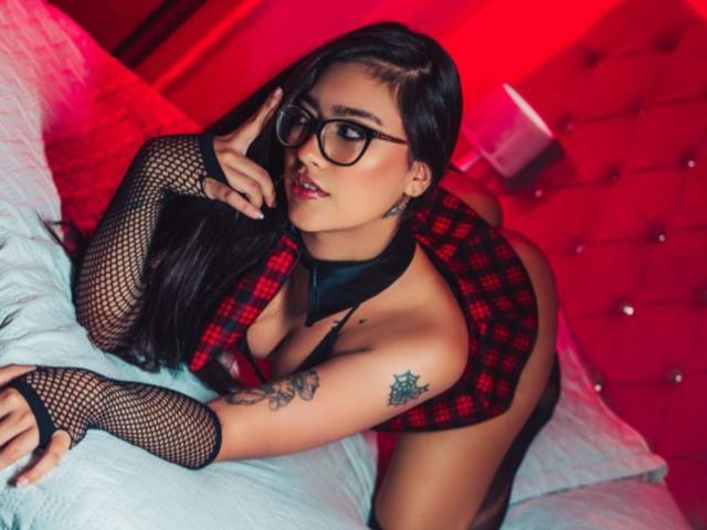 AgathaHowlandd with black glasses, fishnet sleeves, red plaid top, black choker, and tattoos, on a bed, looking seductively at the camera, with red lighting.