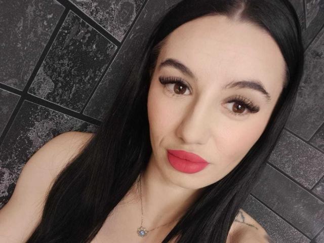 BriannaBratty with long black hair, red lipstick, and brown eyes, wearing a necklace with a small pendant, has a tattoo on her right shoulder, looking at the camera.
