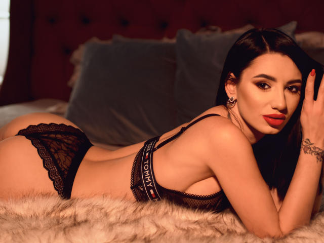 BriannaBratty with long black hair, lying on a furry bed, wearing black lace panties and bra, showing her ass and looking at the camera with red lipstick.