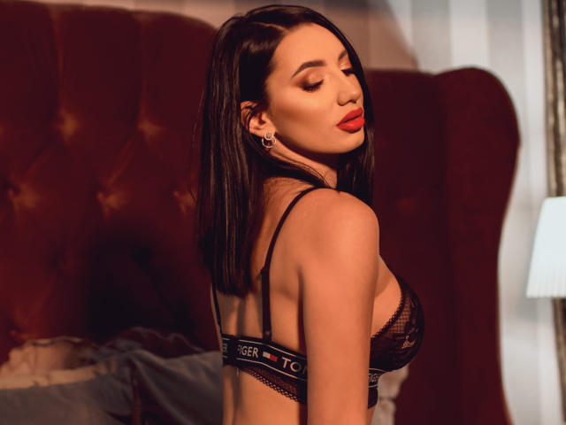 BriannaBratty with red lips, black lace bra, and earrings, standing in front of a brown velvet headboard, showing her tits and back, with closed eyes and sexy pose.