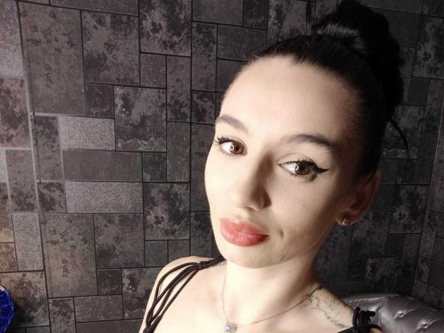 BriannaBratty with dark hair in a bun, wearing black straps, has red lips, long eyelashes, and a slight smirk. Her tits are barely visible, and she's wearing a necklace.