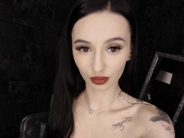 BriannaBratty has pale skin, long black hair, brown eyes, red lipstick, and tattoos on her chest and shoulder. She's wearing a necklace with a small flower.
