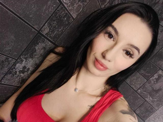 BriannaBratty with long black hair, red tank top, small tits, cute face, light skin, brown eyes, subtle tattoo on right shoulder, necklace, and a gentle smile.