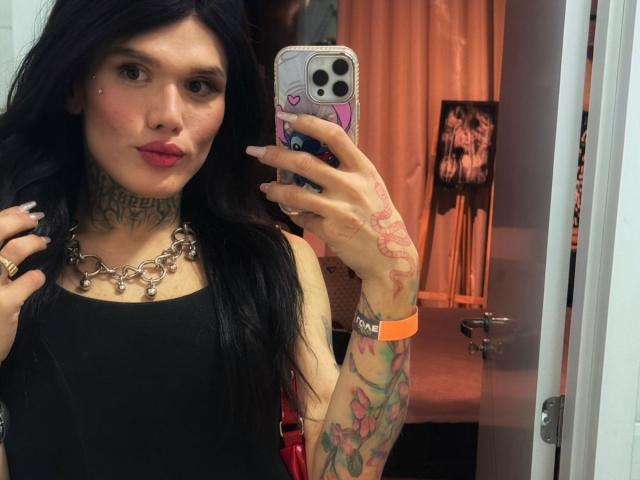 Becab with long black hair, tattoos on neck and arm, wearing a black tank top and chunky necklace, taking a mirror selfie with a pink phone case.