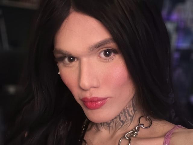 Becab has long black hair, red lipstick, a tattooed neck, and a silver chain collar, with a serious expression and a small nose piercing.