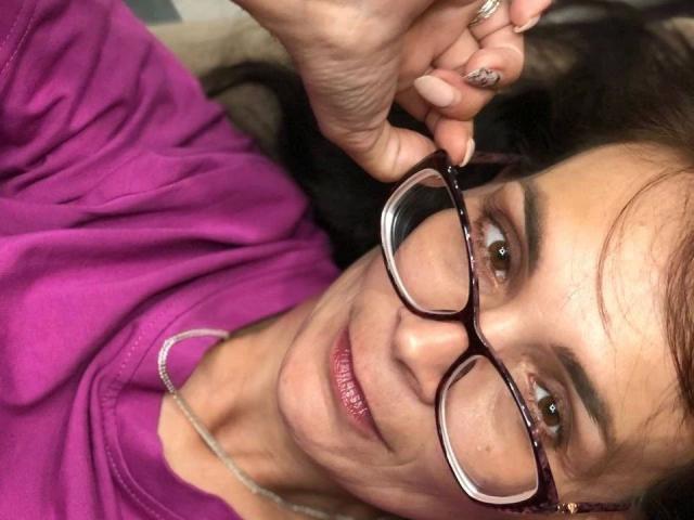 DarkOpiumX-hot is lying down, wearing purple clothes, holding up her glasses with a ringed hand, and has a subtle smile with pink lipstick. Her nails are painted.