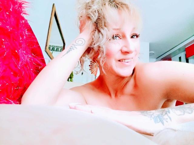 SammyDesire with blonde hair, red polka-dot bra, small tits, tattoo on left wrist, hand near mouth, sitting on beige couch, red pillow in background, smiling at camera.