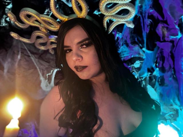 NathalyRose69 with dark makeup, black lipstick, long wavy hair, bare shoulders, gold snake headpiece, and blue light, looking seductively at camera, candle glow in background.