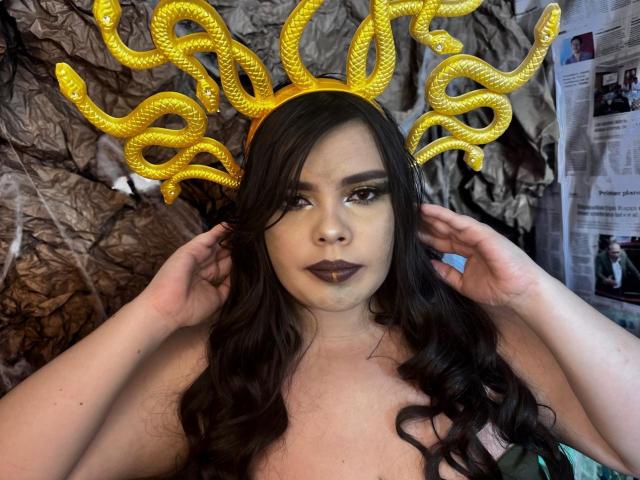 NathalyRose69 with long dark hair, wearing a golden snakehead crown, black lipstick, and touching her hair, showing her bare boobs, in a photo with a crumpled brown background.