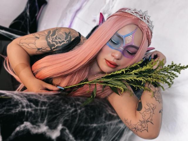 DominicTheOne with pink hair, purple mask, and devil horns, holding greenery, tattooed arms, sleeping on black web-covered surface, wearing silver tiara, pink lips, and blue nail polish.