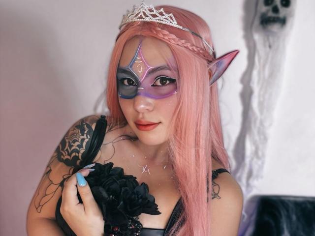 DominicTheOne with pink hair, elf ears, purple mask, tiara, black lace top, holding fan, tattoos on shoulder, blue nail, skull on wall, looking at camera.