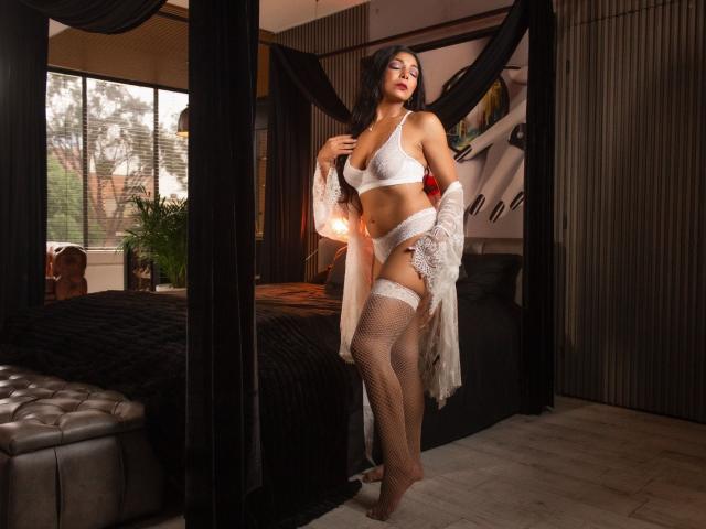 VenusChaud stands in a bedroom wearing white lace lingerie, fishnet stockings, and a sheer robe, exposing her tits and pussy, with her ass slightly visible, looking seductive.