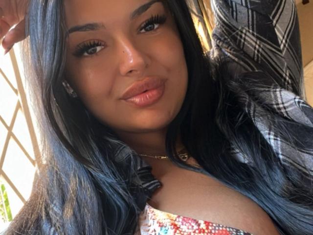 HotKarma with long black hair, full eyebrows, and glossy pink lips, wearing a plaid shirt and a colorful top, showing her cleavage and a subtle smile.
