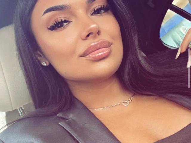 HotKarma has glossy brown lips, thick black eyebrows, long black hair, and is wearing a silver necklace with a heart-shaped pendant. Her tits are partially visible.
