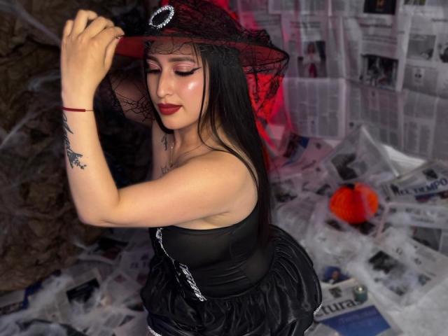 Chloey69 with black hair, wearing a black lace hat, strapless black corset, and black skirt, holding her hat, showing her tits, with a red bracelet and tattoo.