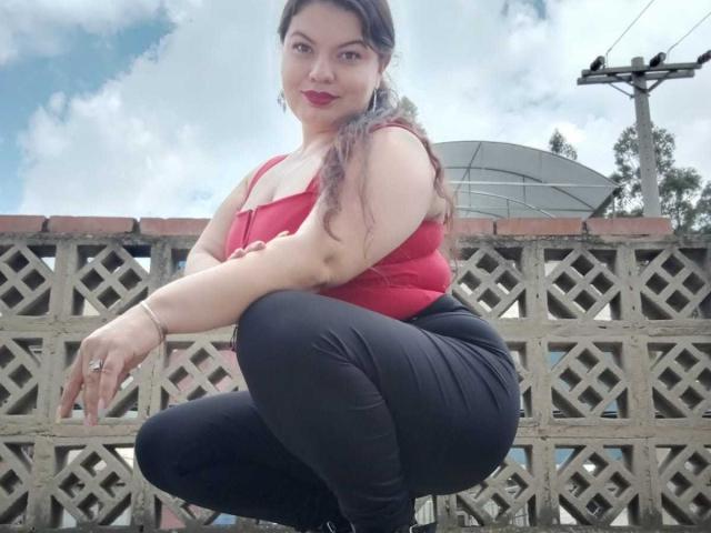 Gaiax69, una donna formosa con lunghi capelli castani, rossetto rosso e una maglia rossa, è accovacciata, indossa pantaloni neri, mostrando il sedere e un anello alla mano sinistra..