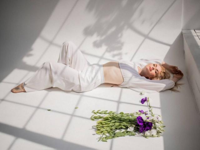 HelloViktoria, lying on white floor, arms above head, wearing white outfit, exposing flat tits, with purple flowers and green stems near her feet, in sunlit room.