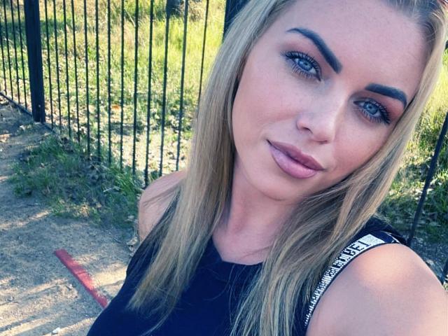 NicoleMelody has blonde hair, blue eyes, and is wearing a black sleeveless top. She has pouty pink lips and a slight smile, with her tits barely visible.