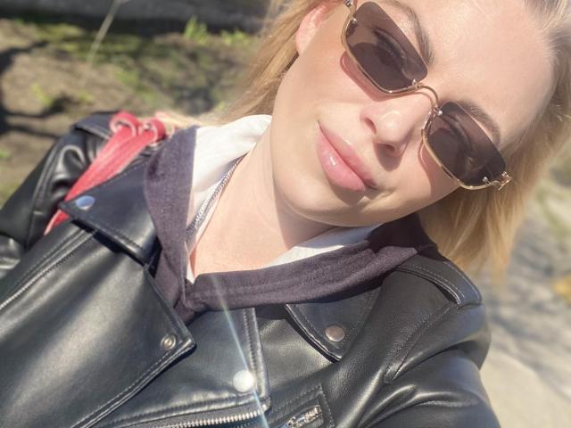 NicoleMelody with blonde hair, wearing aviator sunglasses, black leather jacket, and a dark hoodie, has a sunlit face and slightly parted pink lips, showing a relaxed expression.