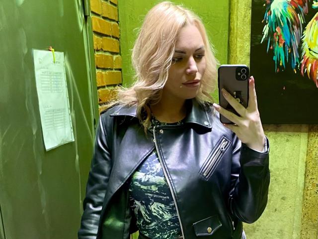 NicoleMelody with blonde hair, wearing a black leather jacket, taking a selfie in a green-walled room, holding a black phone, showing off her tits, and looking cute.