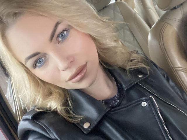 NicoleMelody with blonde hair, blue eyes, and glossy lips, wearing a black leather jacket, sitting in a tan leather car seat. Her tits are subtle, not visible.