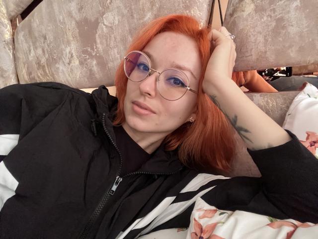 Sarah Rousen mit roten Haaren, Brille, schwarzer Jacke und einem Blumenkissen, die ihren tätowierten Arm und ihre leicht geschwollenen Lippen zeigt, wirkt entspannt. Ihre blauen Augen fallen ins Auge..