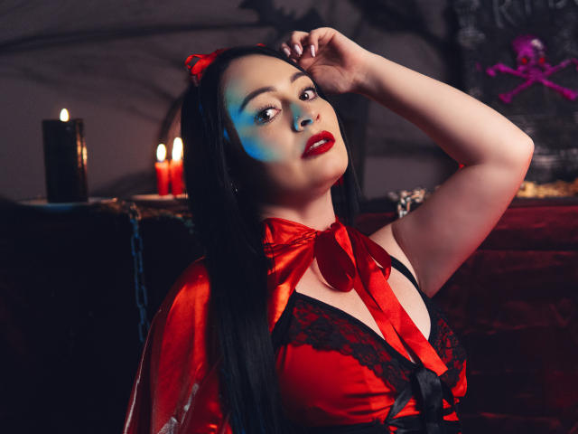 KristelRouse with long black hair, red lipstick, and red satin dress with black lace, showing her big tits, hand in hair, red bow, and red devil horns, candles in background.