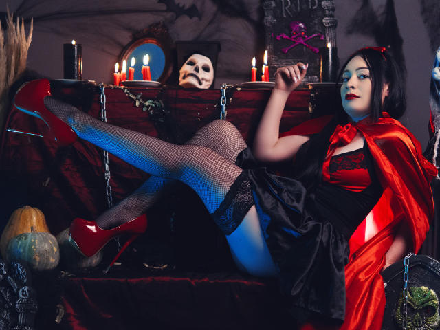 KristelRouse with red high heels, black fishnet stockings, red satin cape, black dress, and red lipstick, reclining on a dark velvet sofa with lit candles and a skull.