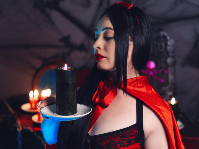 KristelRouse with black lace top, red cape, and red lipstick, holding a lit black candle on a plate, looking to the side, with candles in the background.