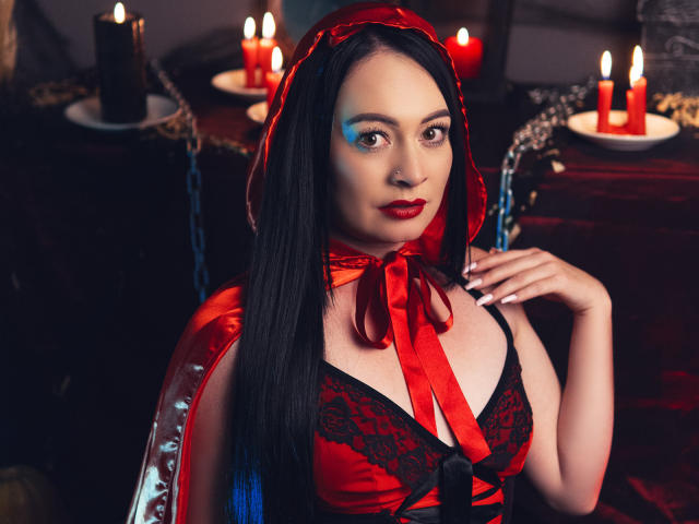 KristelRouse with long black hair, red hooded dress, black lace, red ribbon, red lipstick, sitting, candles in background, looking at camera, hand on chest, sexy, seductive.