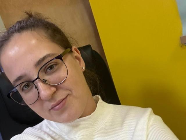 BellGabi with glasses, brown hair in a bun, white turtleneck, small earrings, slightly smiling, yellow wall in background, black chair behind. Soft skin, light makeup.