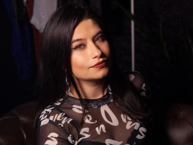 MarianaConti with dark hair, brown eyes, and red lipstick, wearing a sheer black top with white swirls, showing her tits. She's looking up with a slight smile.