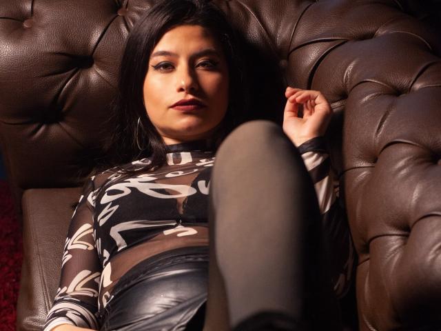 MarianaConti lying on a brown leather couch, wearing a sheer black top with white patterns, black leather pants, and black stockings, with a relaxed, slightly seductive expression.